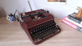 A serviced Olympia SM typewriter in dark burgundy red sits on a light wood desk, from three-quarter front view