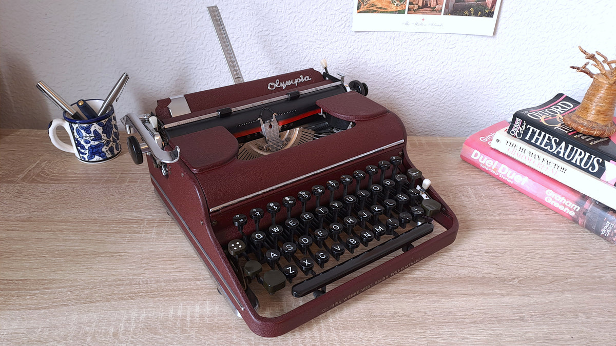 A serviced Olympia SM typewriter in dark burgundy red sits on a light wood desk, from three-quarter front view